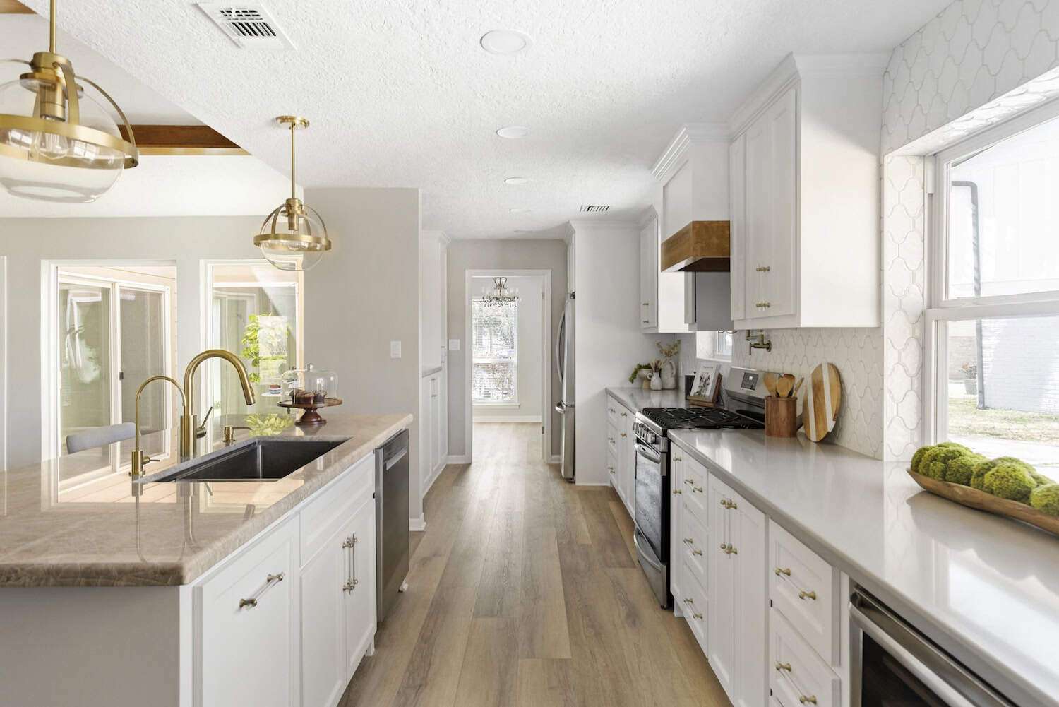Bright galley-style kitchen featuring white cabinets, gold pendant lights, wood flooring, marble countertops, and stainless steel appliances.
