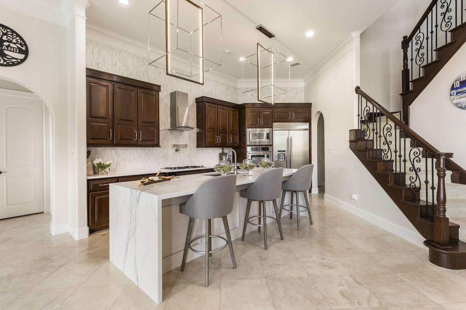 Elegant kitchen featuring dark wood cabinetry, marble waterfall island with seating, stainless steel appliances, and open staircase with wrought iron railing.