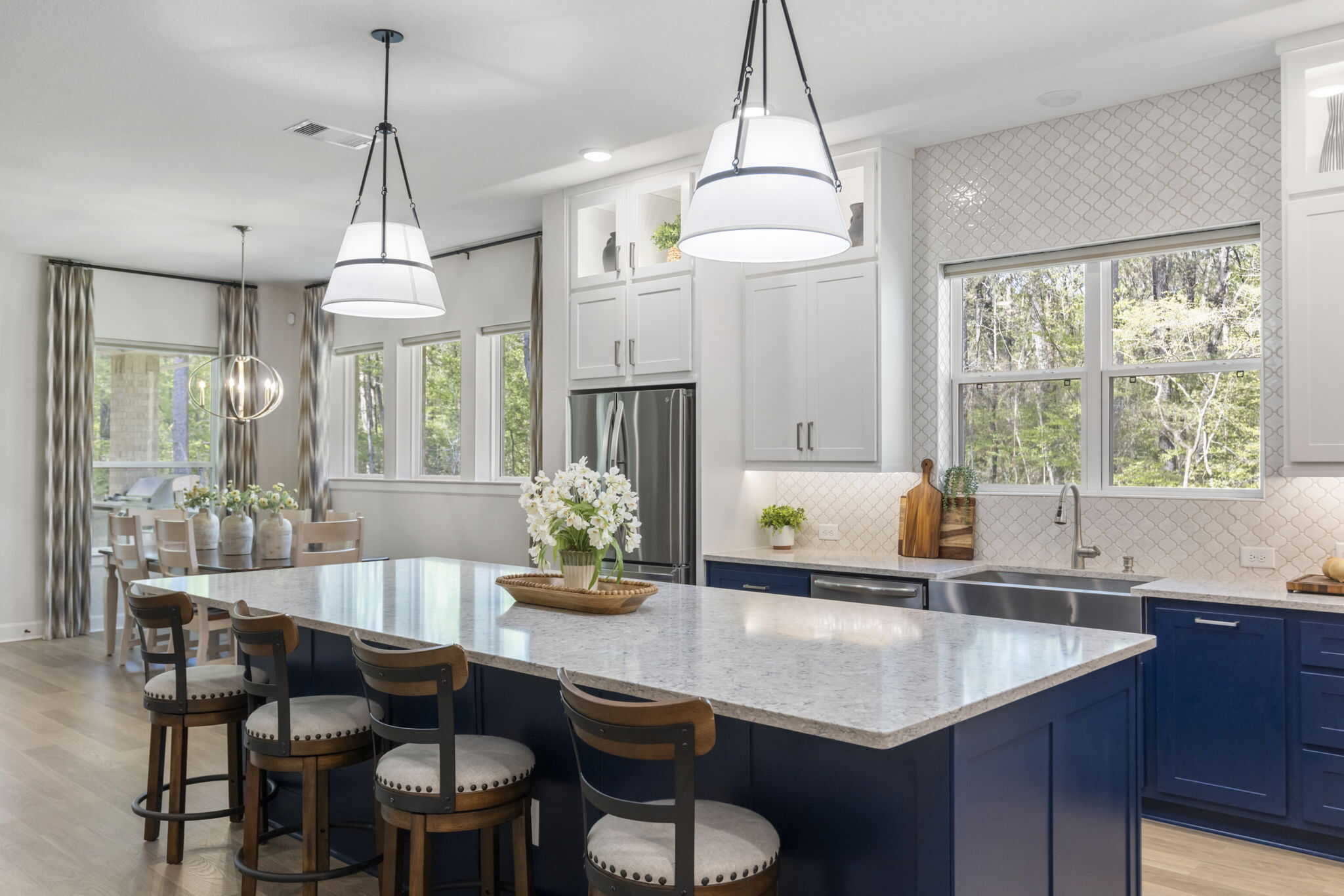 Bright modern kitchen featuring a large navy blue island with seating, white upper cabinets, pendant lights, stainless steel appliances, and wood flooring.