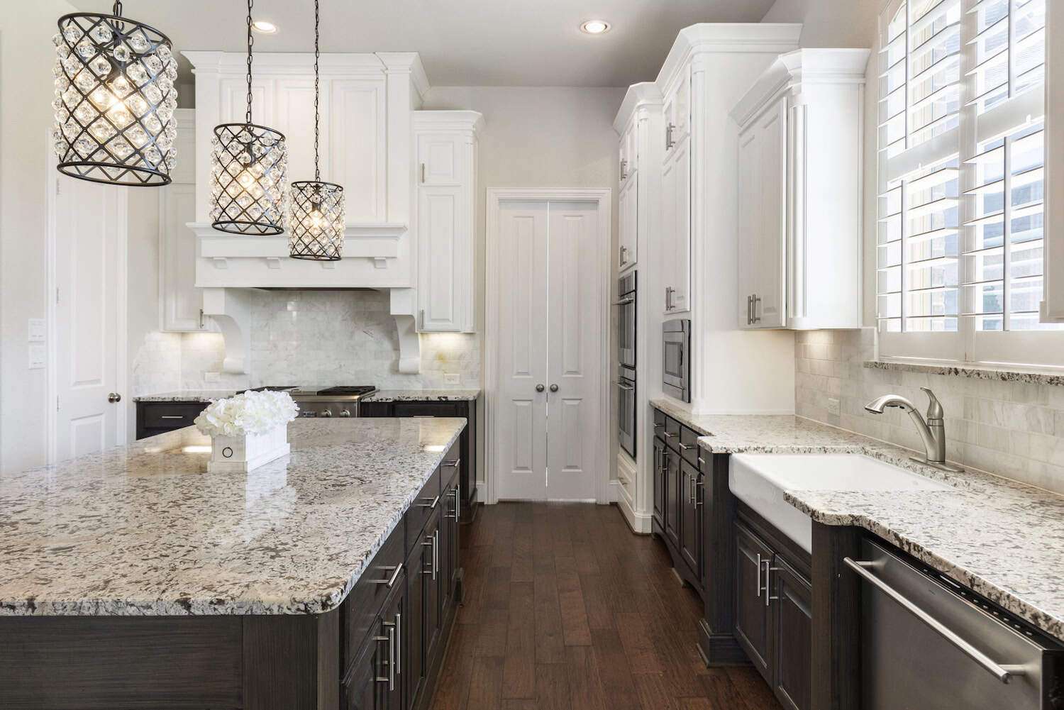 Bright kitchen with granite countertops and pendant lighting Elegant kitchen with white upper cabinets, dark lower cabinets, granite countertops, farmhouse sink, crystal pendant lights, and hardwood flooring