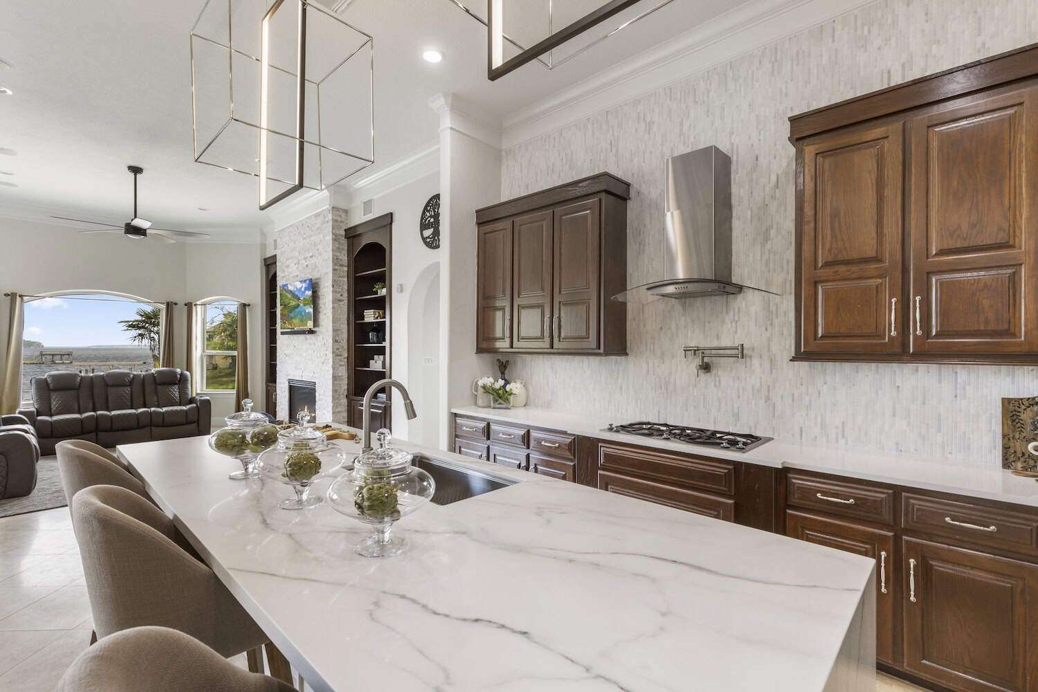 Modern kitchen with marble island, dark wood cabinets, and open view of living room with stone fireplace.