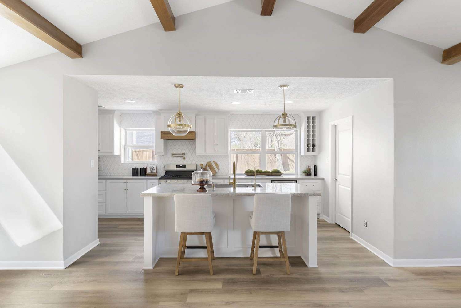 Modern white kitchen with wooden ceiling beams, island with bar stools, and pendant lighting.