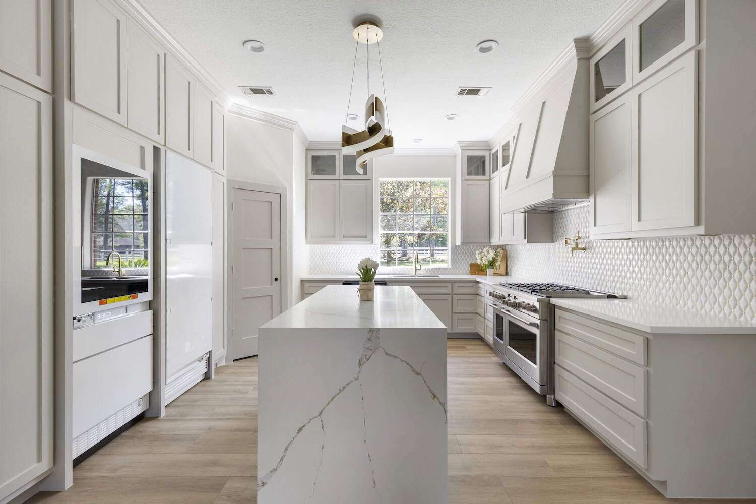 Contemporary all-white kitchen with sleek cabinets, marble waterfall island, geometric pendant light, and large window.