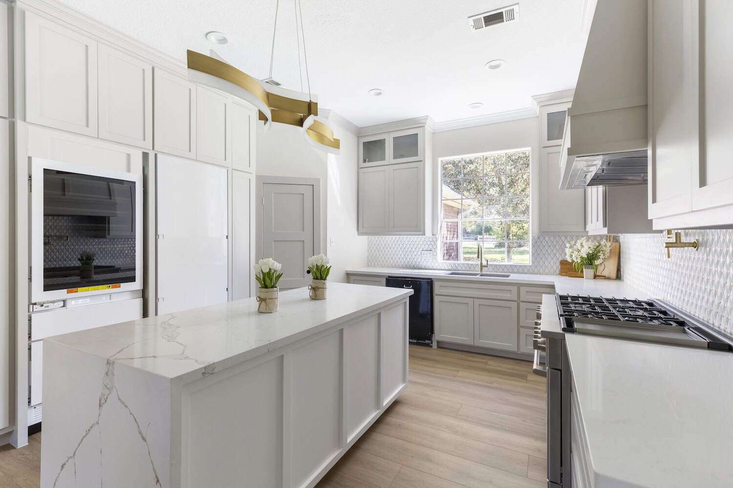 Elegant white kitchen with marble island Sleek white kitchen featuring a large marble island, gold modern chandelier, light wood flooring, and stainless steel appliances.