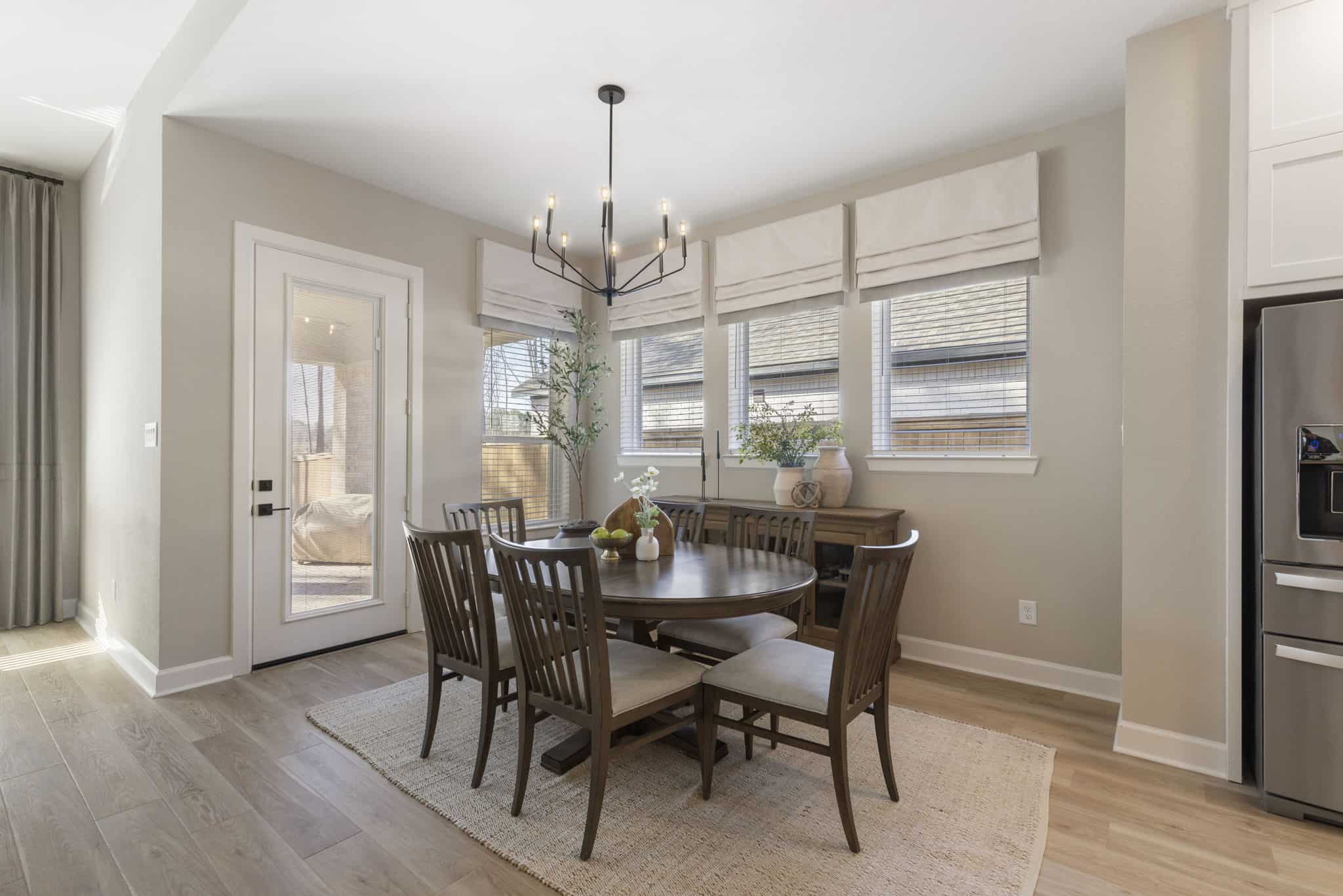 Modern dining room with natural light and wooden furniture Bright dining area with round wooden table, modern chandelier, and large windows with Roman shades.