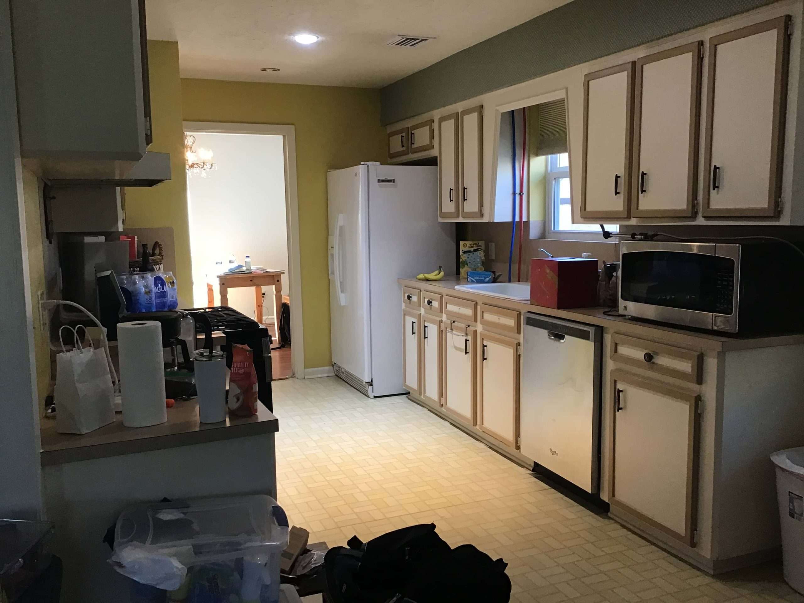 Kitchen before renovation with older cabinets, laminate flooring, white appliances, and cluttered countertops in a traditional layout