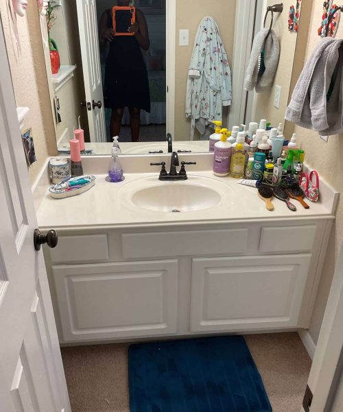 Small bathroom before renovation with a white vanity, mirror, cluttered countertop with toiletries, and beige walls