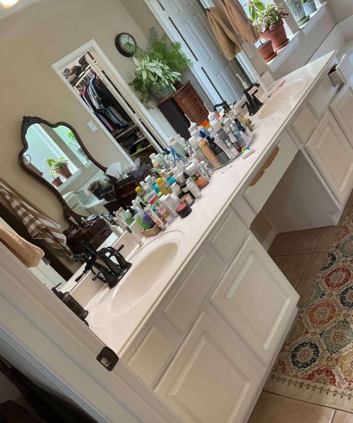 Large bathroom before renovation with double sink vanity, mirror, and cluttered countertop full of toiletries and personal items
