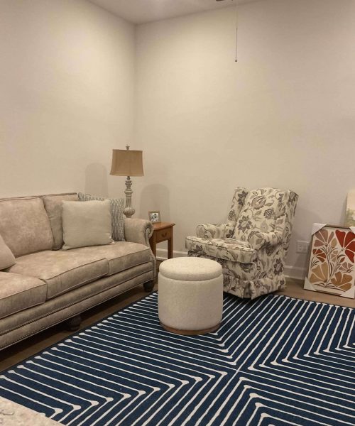 Before Renovation – Living Room with Neutral Furniture and Blue Rug Living room before renovation with beige sofa, patterned armchair, ottoman, and blue geometric rug