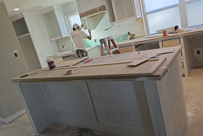 Kitchen renovation in progress with new cabinets being installed and painted, island under construction, and contractor working on backsplash area