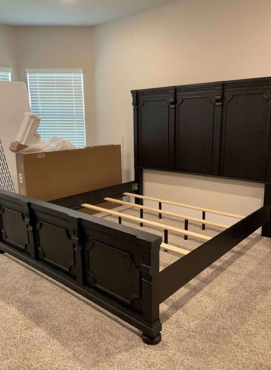 Before Renovation – Bedroom with Black Wood Bed Frame Bedroom before renovation with black wood bed frame being assembled, visible box packaging, and neutral carpet