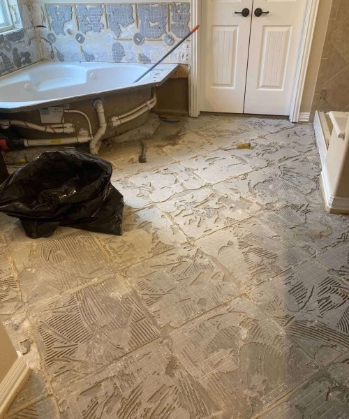 Bathroom during renovation with old tile flooring removed, adhesive residue on the subfloor, and bathtub plumbing area exposed