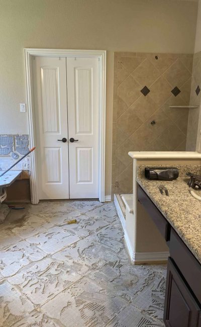 Bathroom renovation in progress with removed flooring, exposed subfloor, partially removed vanity area, and shower visible in the background