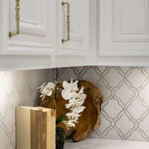 Close-up of white kitchen cabinets with brass handles, decorative tile backsplash, and styled countertop with books and orchid