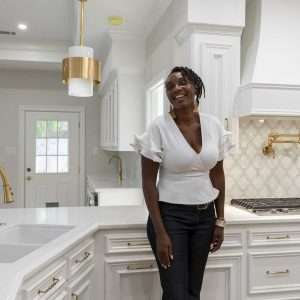 Woman standing in a bright white kitchen with gold fixtures, pendant lighting, and decorative tile backsplash
